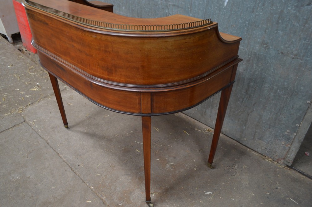 An Edwardian mahogany Carlton House desk featuring a raised U-shaped gallery of drawers, a brass rail, a green leather writing surface, and tapered square legs with castors. View IV