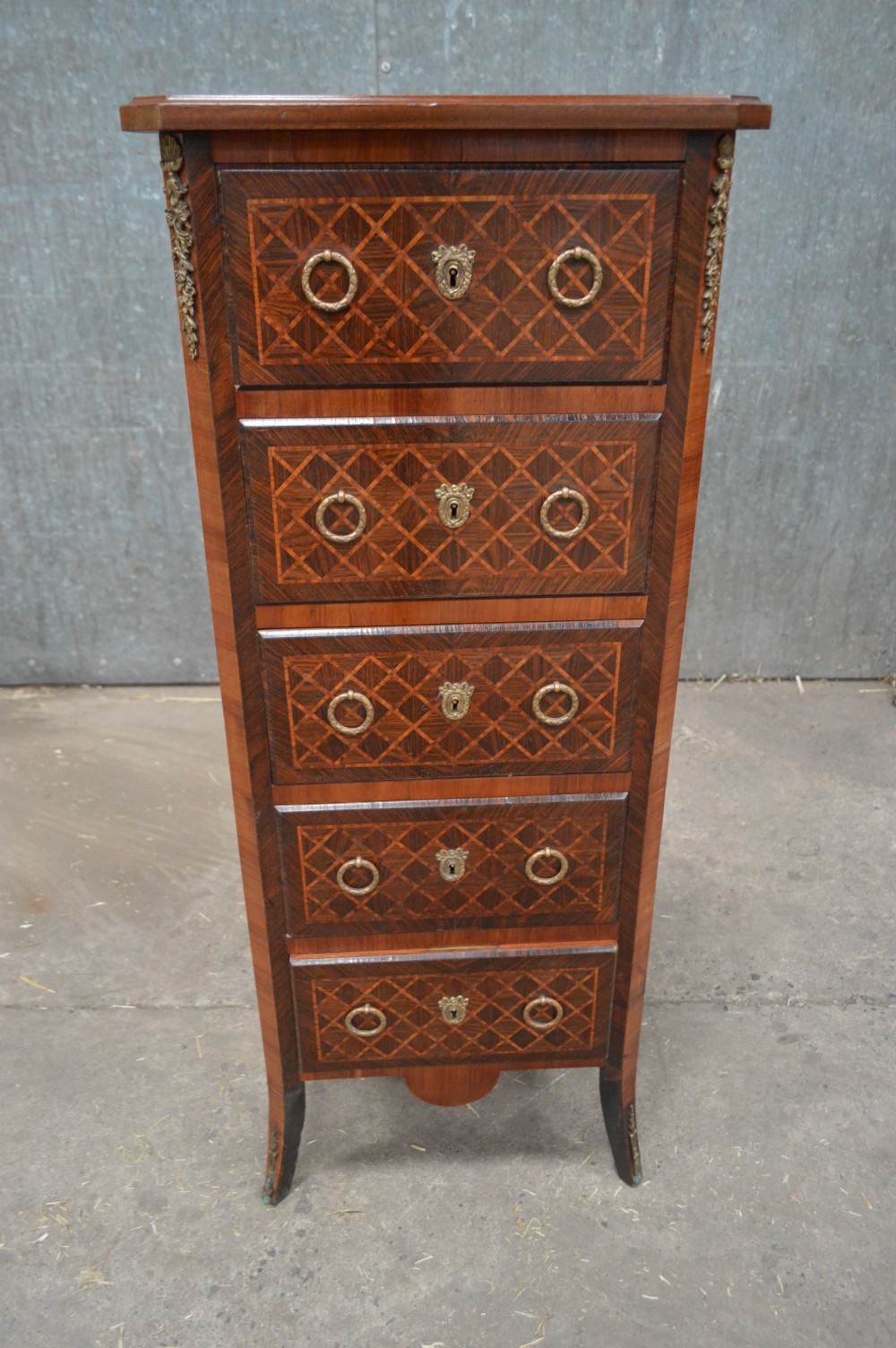 A tall, narrow 19th-century French timber chest featuring five drawers with geometric diamond parquetry inlay, brass ring handles, ornate keyhole escutcheons, and gilded corner mounts.