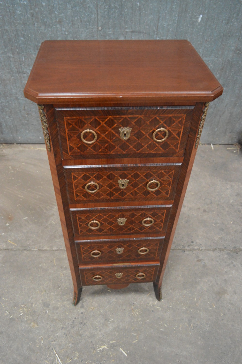 A tall, narrow 19th-century French timber chest featuring five drawers with geometric diamond parquetry inlay, brass ring handles, ornate keyhole escutcheons, and gilded corner mounts. View II