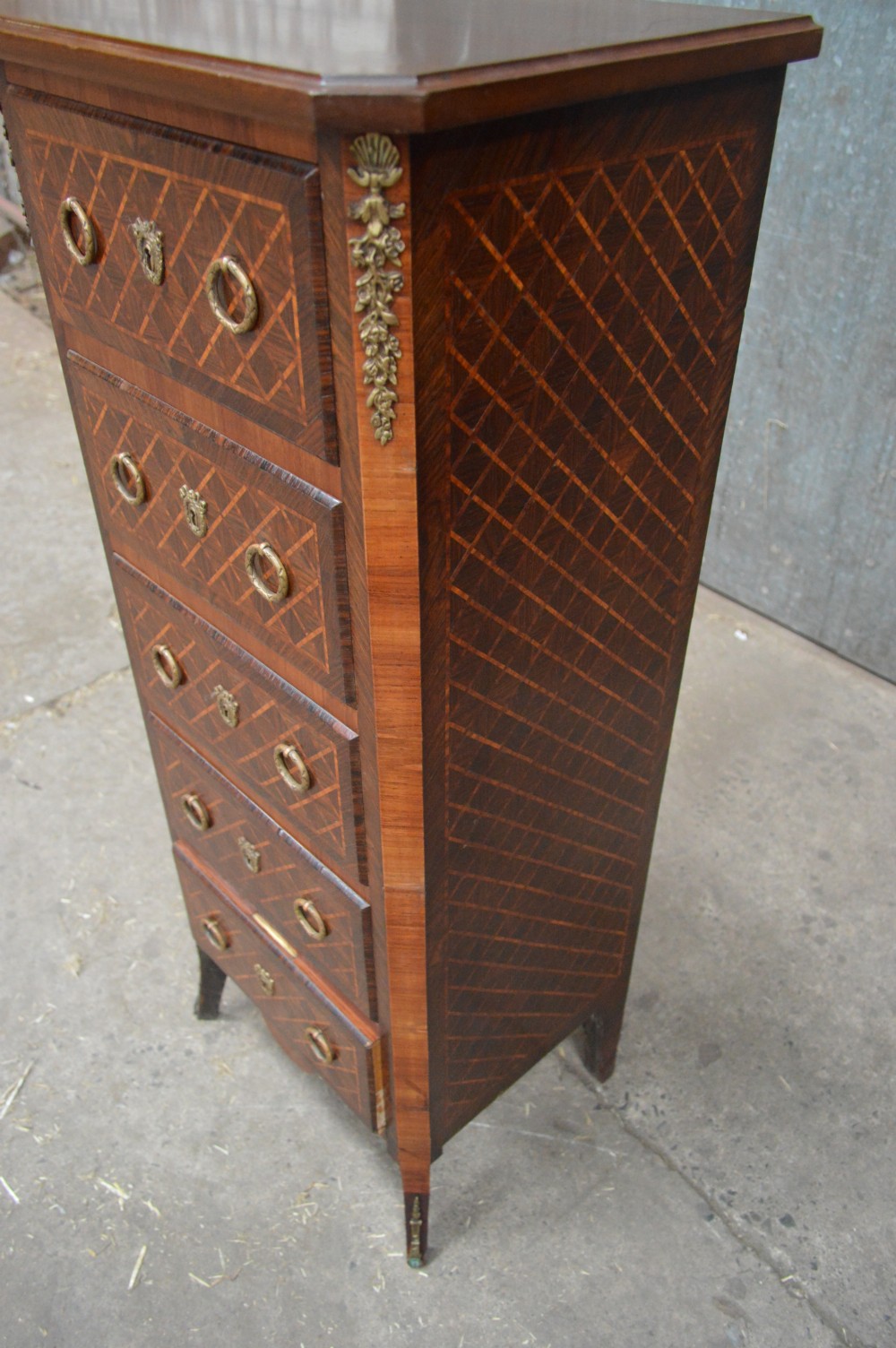 A tall, narrow 19th-century French timber chest featuring five drawers with geometric diamond parquetry inlay, brass ring handles, ornate keyhole escutcheons, and gilded corner mounts. View IV
