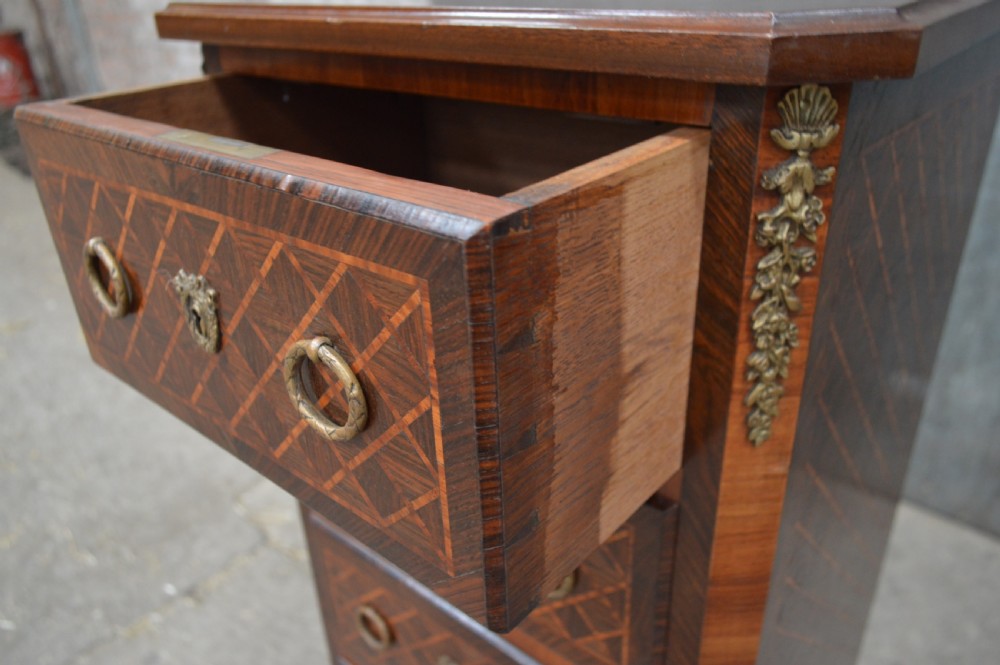 A tall, narrow 19th-century French timber chest featuring five drawers with geometric diamond parquetry inlay, brass ring handles, ornate keyhole escutcheons, and gilded corner mounts. View V