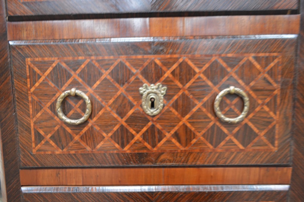 A tall, narrow 19th-century French timber chest featuring five drawers with geometric diamond parquetry inlay, brass ring handles, ornate keyhole escutcheons, and gilded corner mounts. View VI