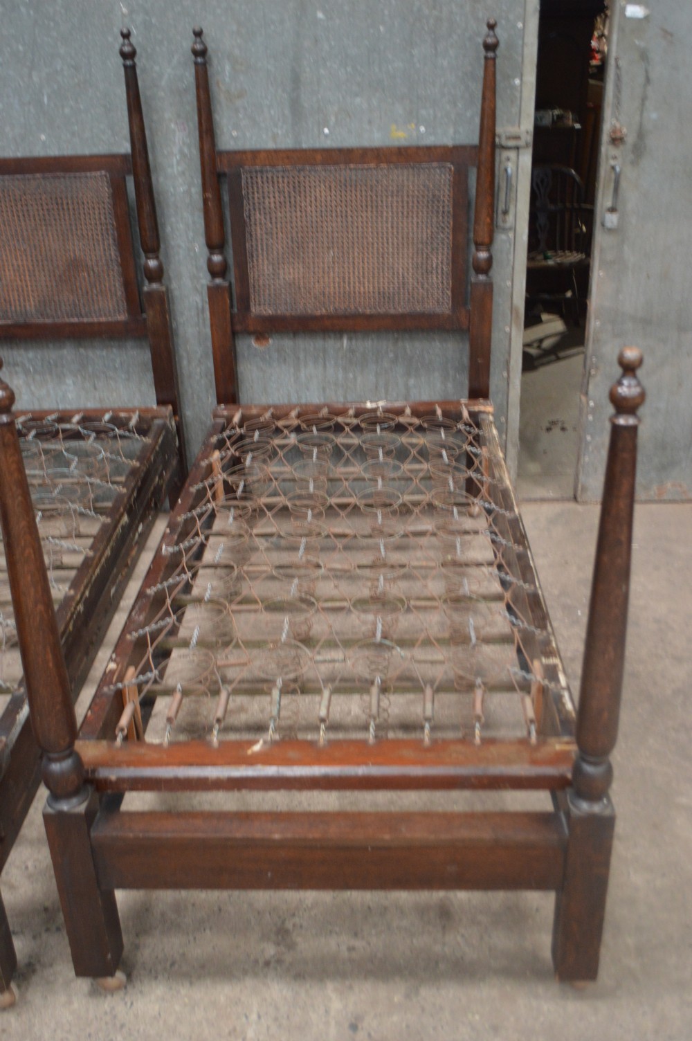 A matching pair of dark oak single bed frames featuring woven cane headboards, turned corner posts with finials, and castor wheels. View V