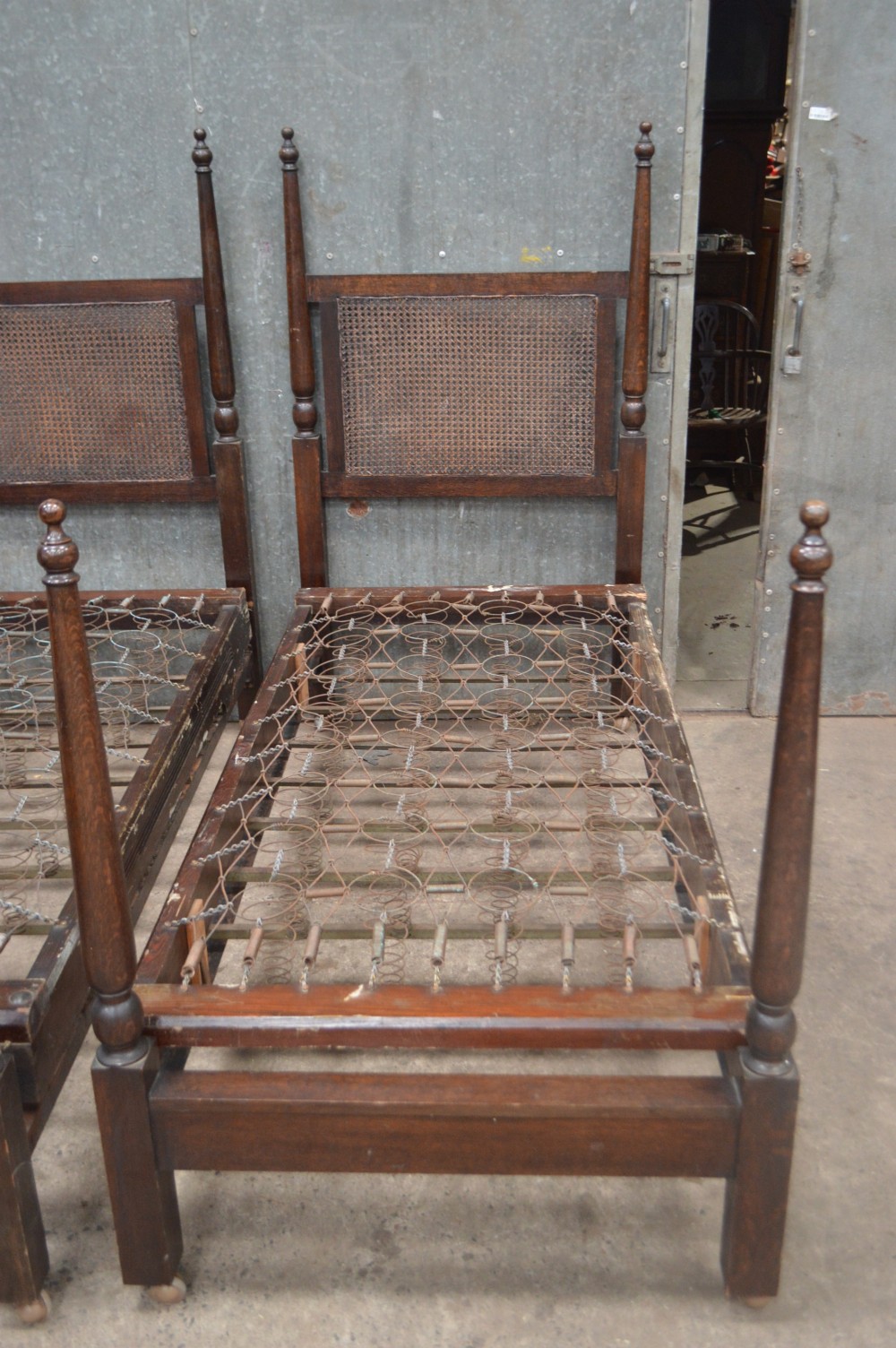 A matching pair of dark oak single bed frames featuring woven cane headboards, turned corner posts with finials, and castor wheels. View VI