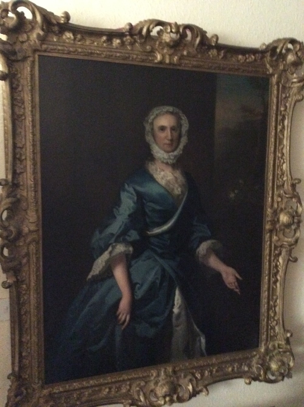 An 18th-century oil painting on canvas depicting a seated woman in a blue satin dress and lace cap, displayed inside an ornate carved giltwood frame. View IV
