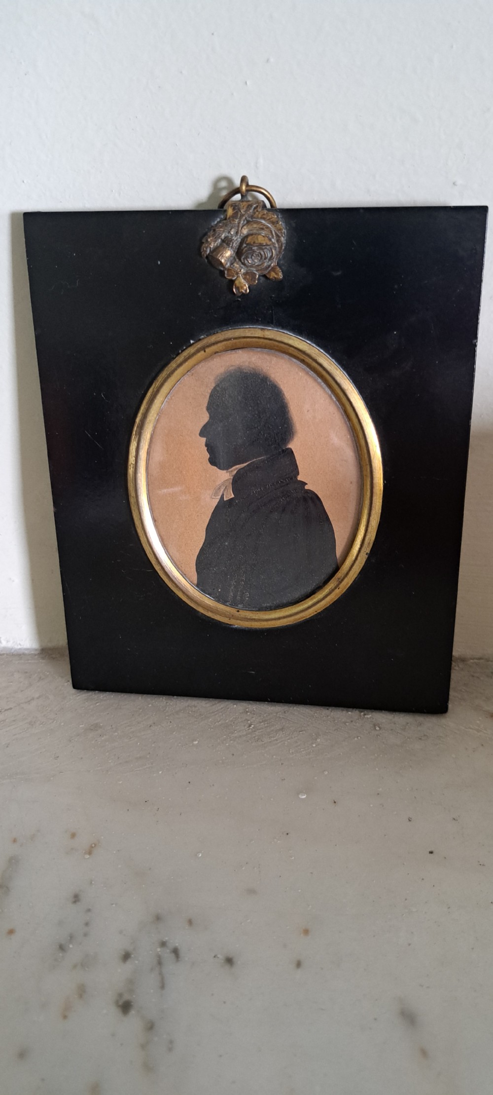 A profile silhouette portrait of a 19th-century gentleman set within a gilded oval bezel mounted inside a square black frame with a brass acorn hanger.