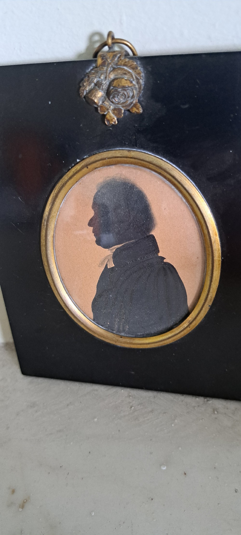 A profile silhouette portrait of a 19th-century gentleman set within a gilded oval bezel mounted inside a square black frame with a brass acorn hanger. View II