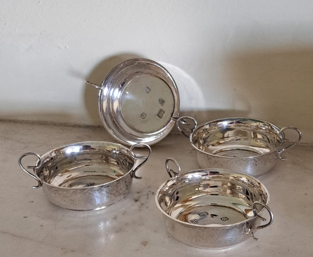 A set of four small, round sterling silver wine tasters with twin loop handles and visible hallmarks stamped on the polished interiors.