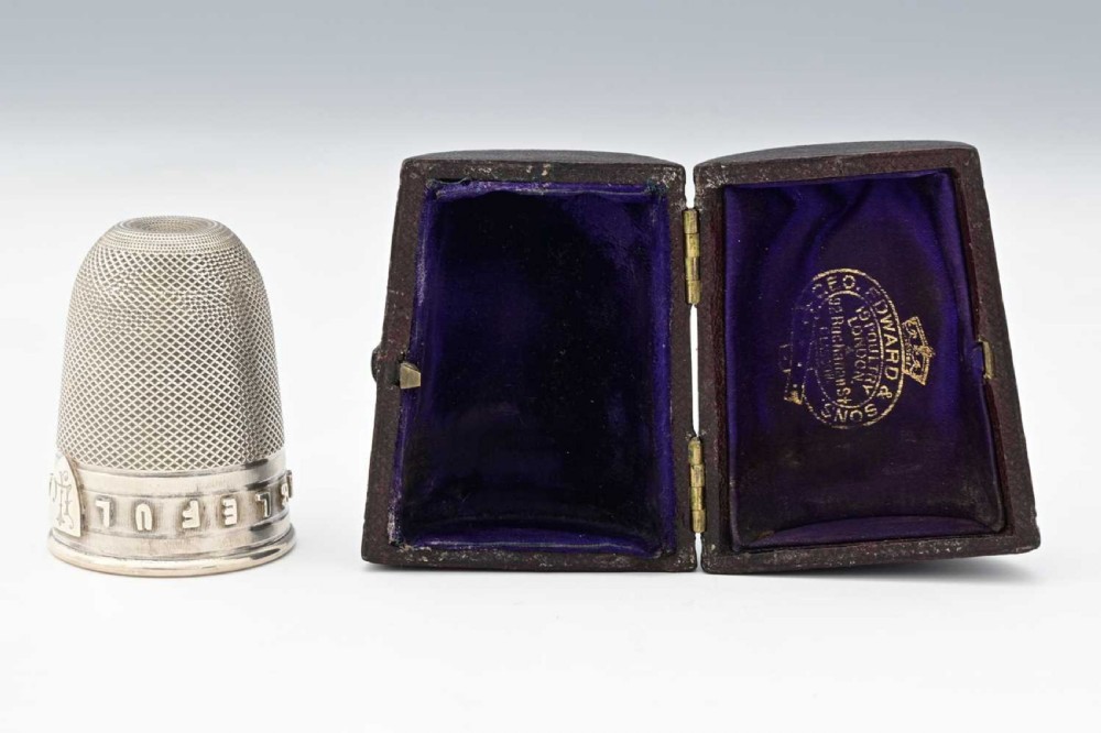 A Victorian sterling silver novelty spirit cup shaped as an oversized thimble with engine-turned texturing and raised lettering reading JUST A THIMBLEFUL, presented next to its plush-lined leather case.