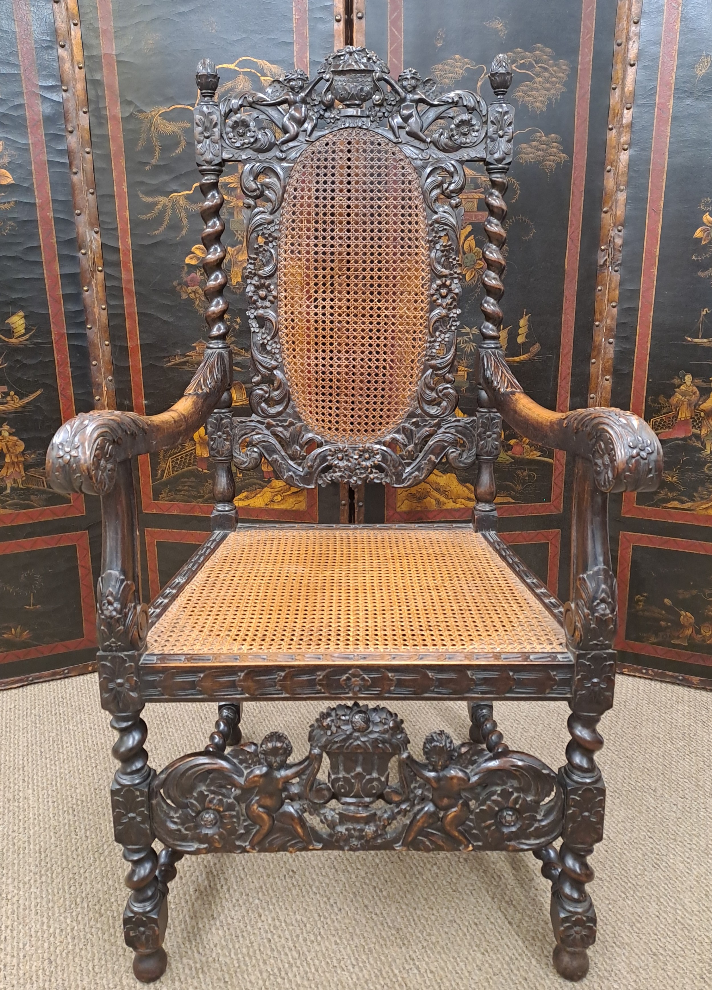 A dark carved oak armchair featuring a woven cane backrest and seat frame, flanked by barley twist uprights and intricate carvings of cherubs.