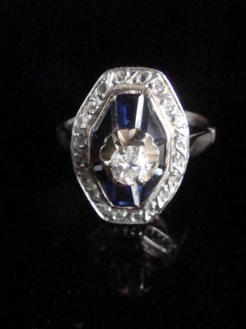 fig 1. An Art Deco 18ct white gold ring featuring a central diamond encircled by dark blue calibrated sapphires and an outer geometric diamond bezel.