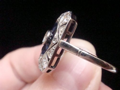 fig 1. An Art Deco rectangular white gold ring featuring a central round diamond surrounded by calibre-cut blue sapphires and framed by smaller diamonds with detailed millegrain edging.