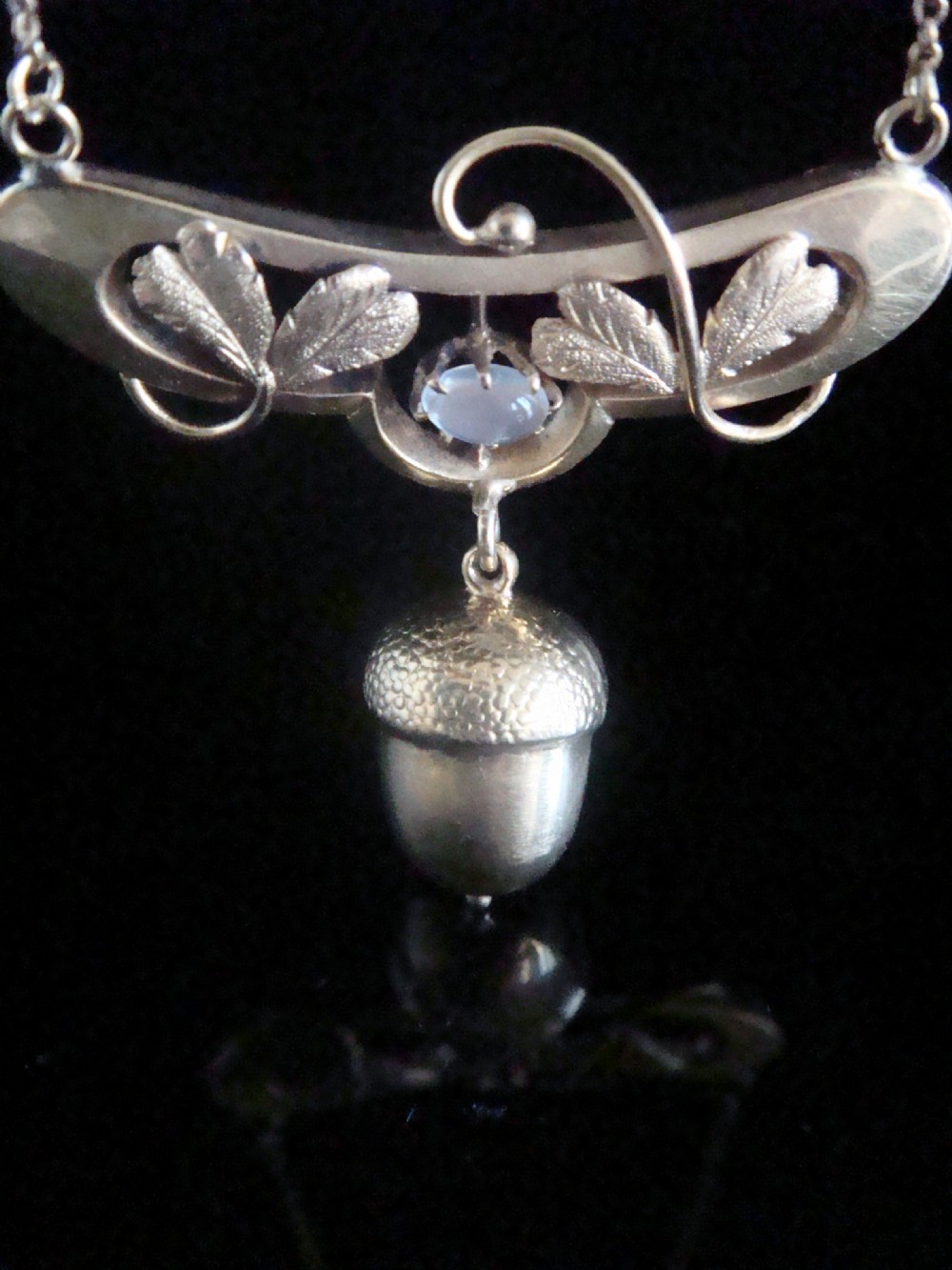 An Art Nouveau yellow gold necklace featuring a central oval moonstone, sculpted leaves, and a textured hanging acorn pendant.