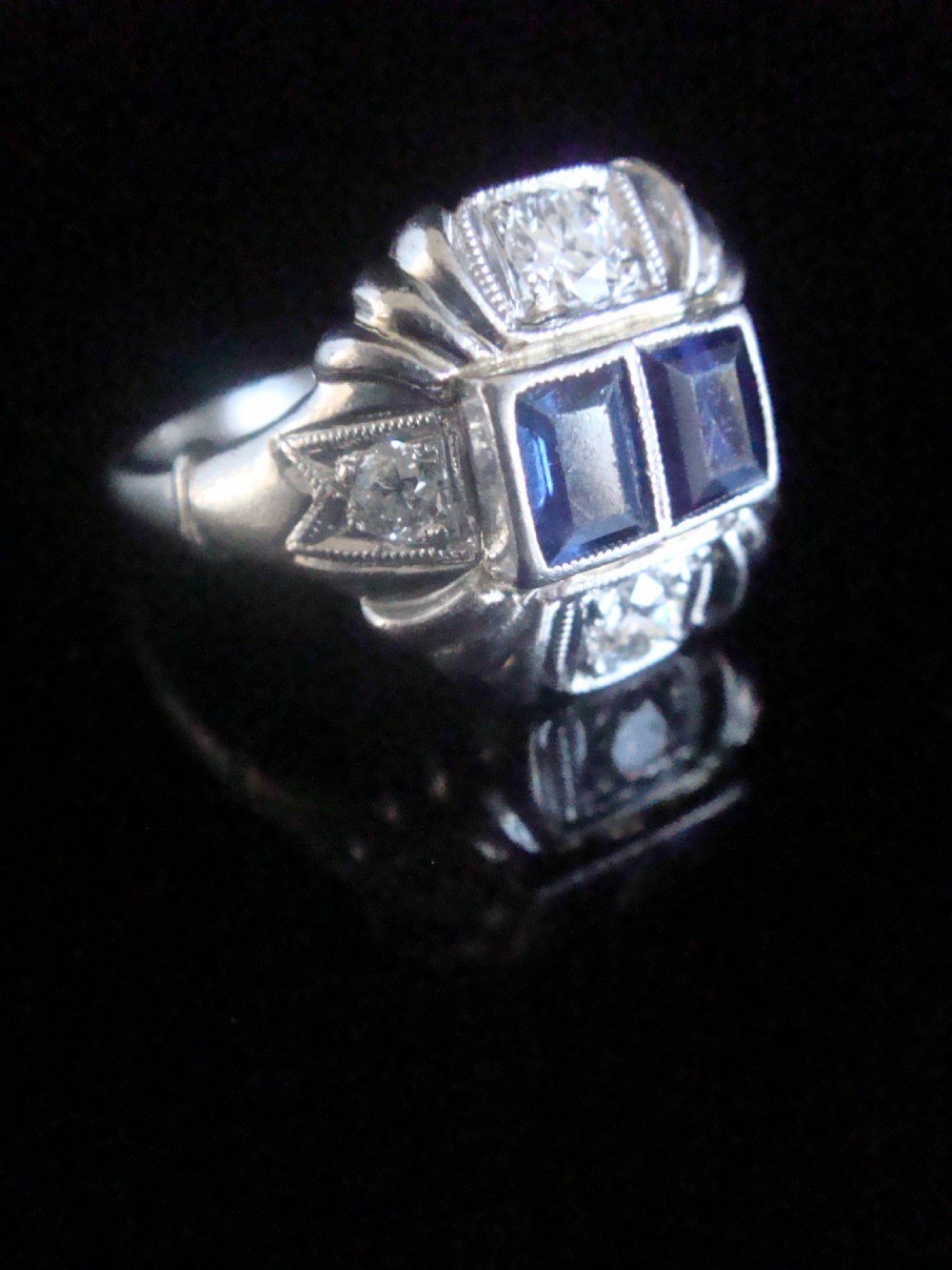 An Art Deco platinum ring featuring two central rectangular blue sapphires flanked by round diamonds and detailed milgrain edging.