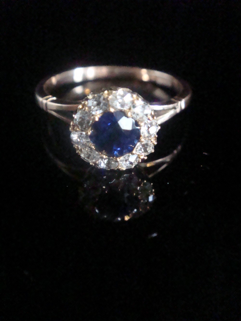 An Edwardian 18ct yellow gold ring with a central round blue sapphire encircled by a halo of old cut diamonds.