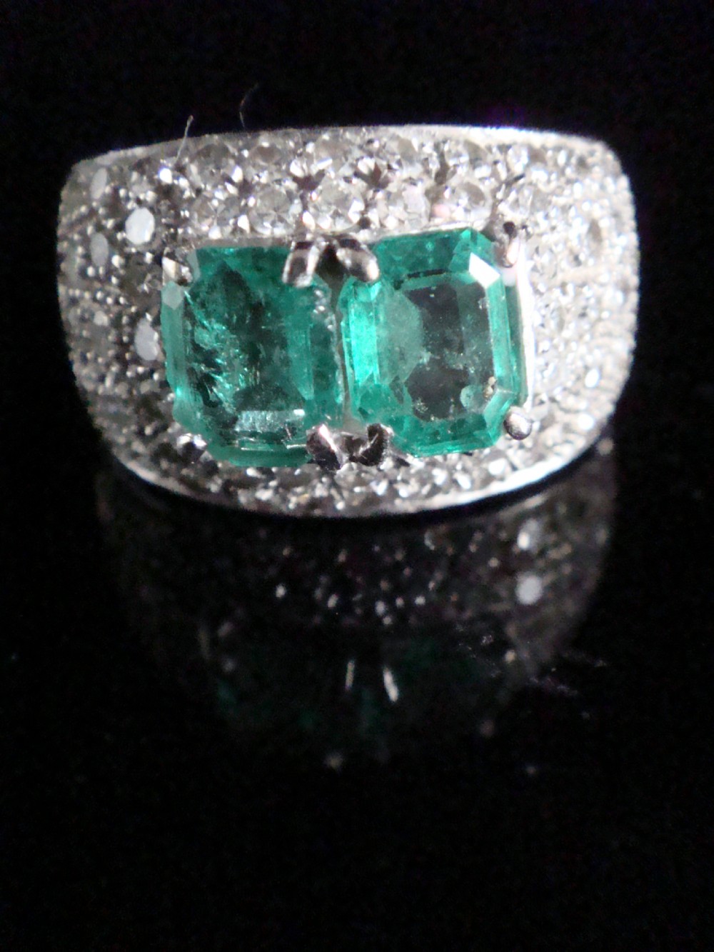 An Art Deco platinum band set with two central emerald-cut green emeralds framed by a broad pave diamond setting.