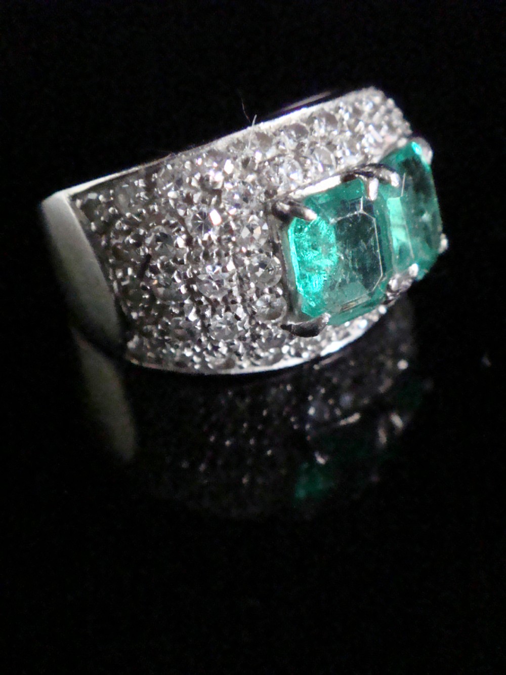 An Art Deco platinum band set with two central emerald-cut green emeralds framed by a broad pave diamond setting. View II