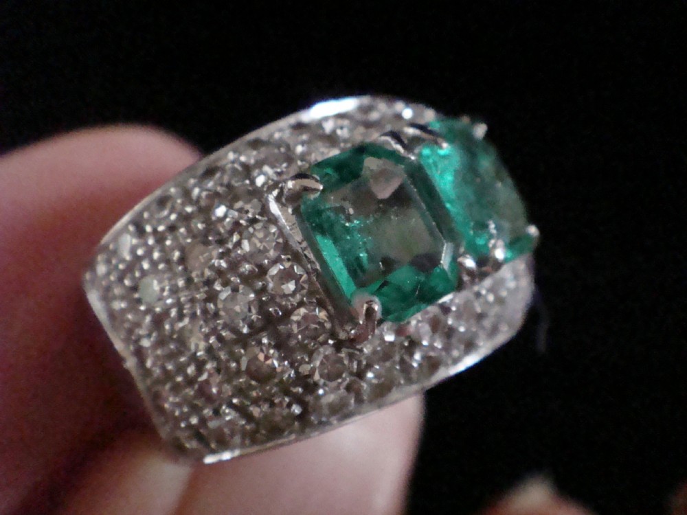 An Art Deco platinum band set with two central emerald-cut green emeralds framed by a broad pave diamond setting. View VI