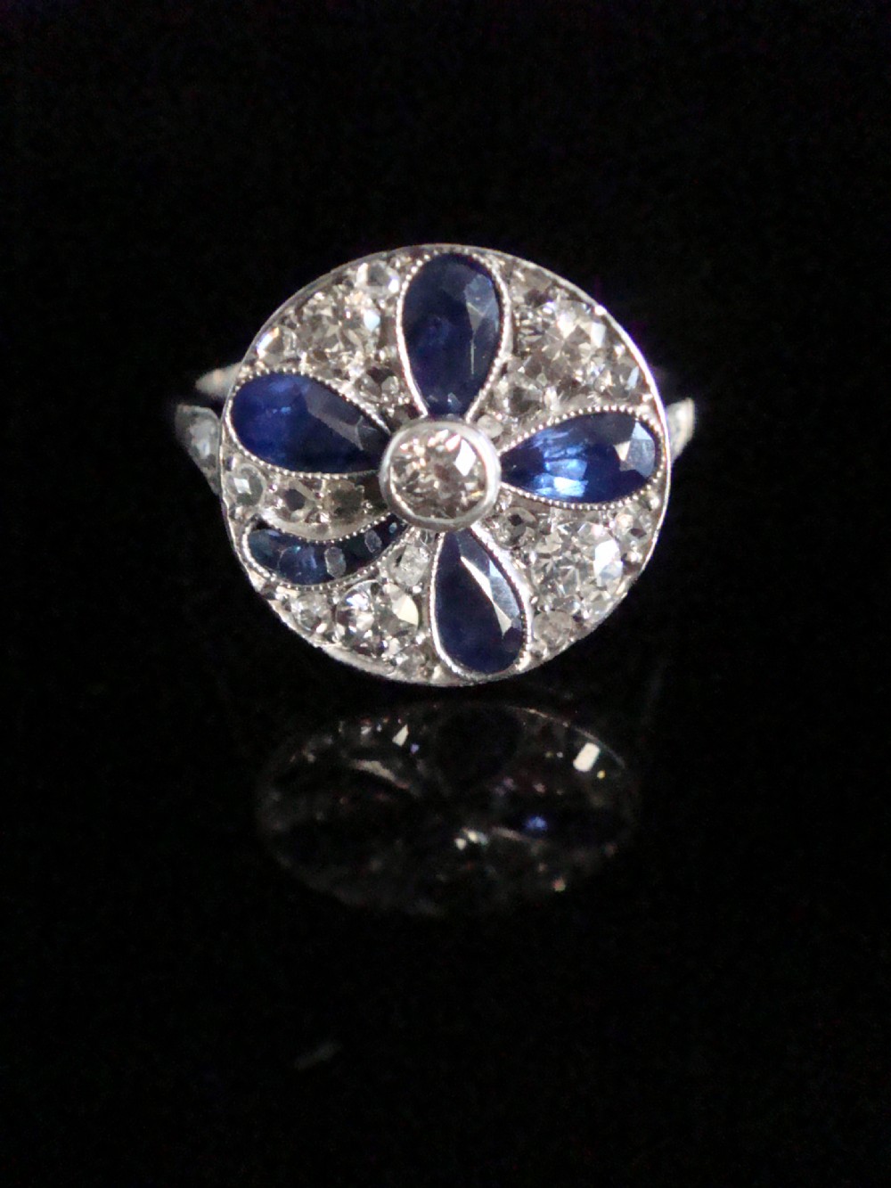 An Art Deco circular platinum ring featuring a central round diamond surrounded by four pear-cut blue sapphires forming a clover motif set amidst pave diamonds.