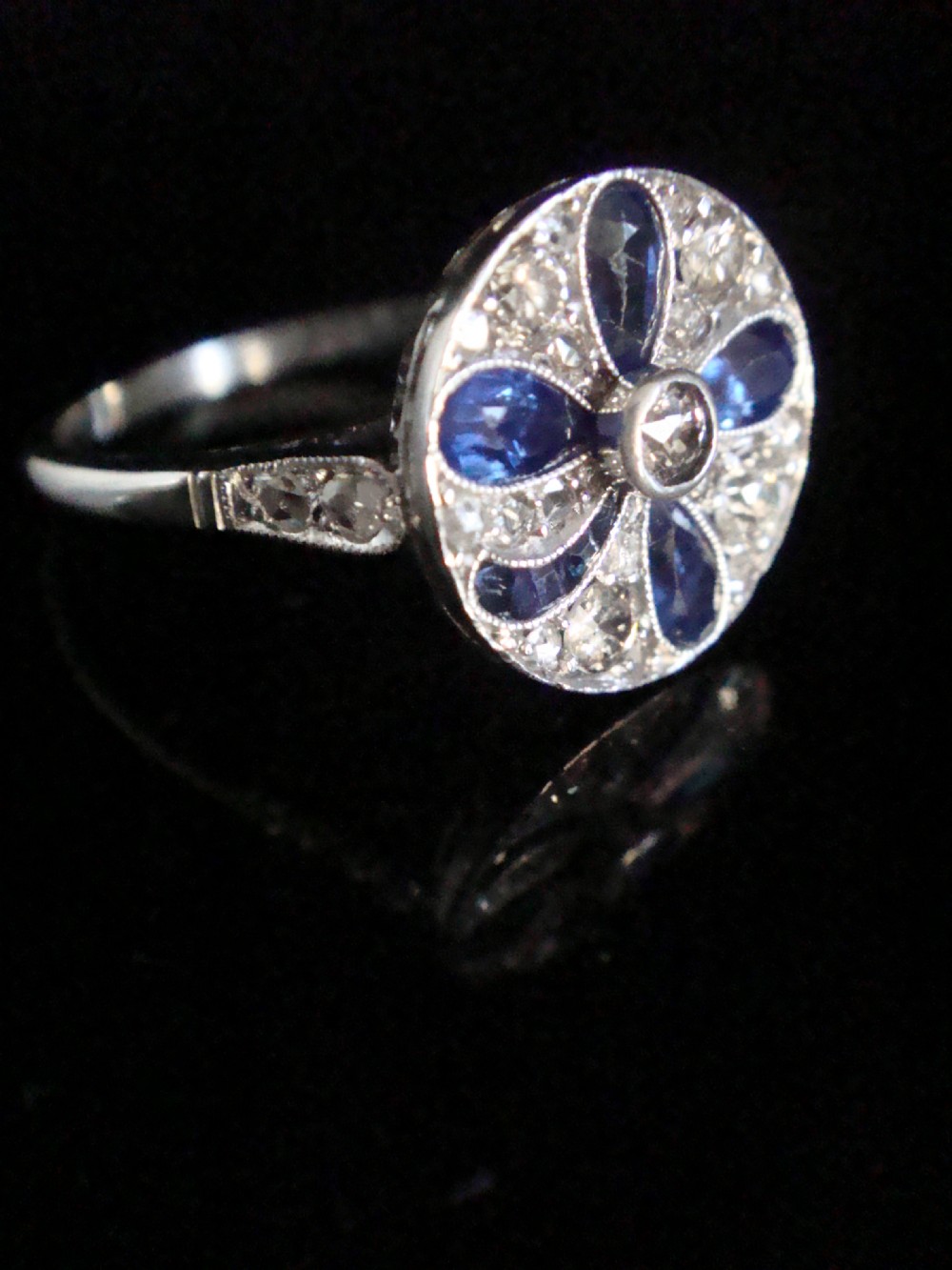 An Art Deco circular platinum ring featuring a central round diamond surrounded by four pear-cut blue sapphires forming a clover motif set amidst pave diamonds. View II