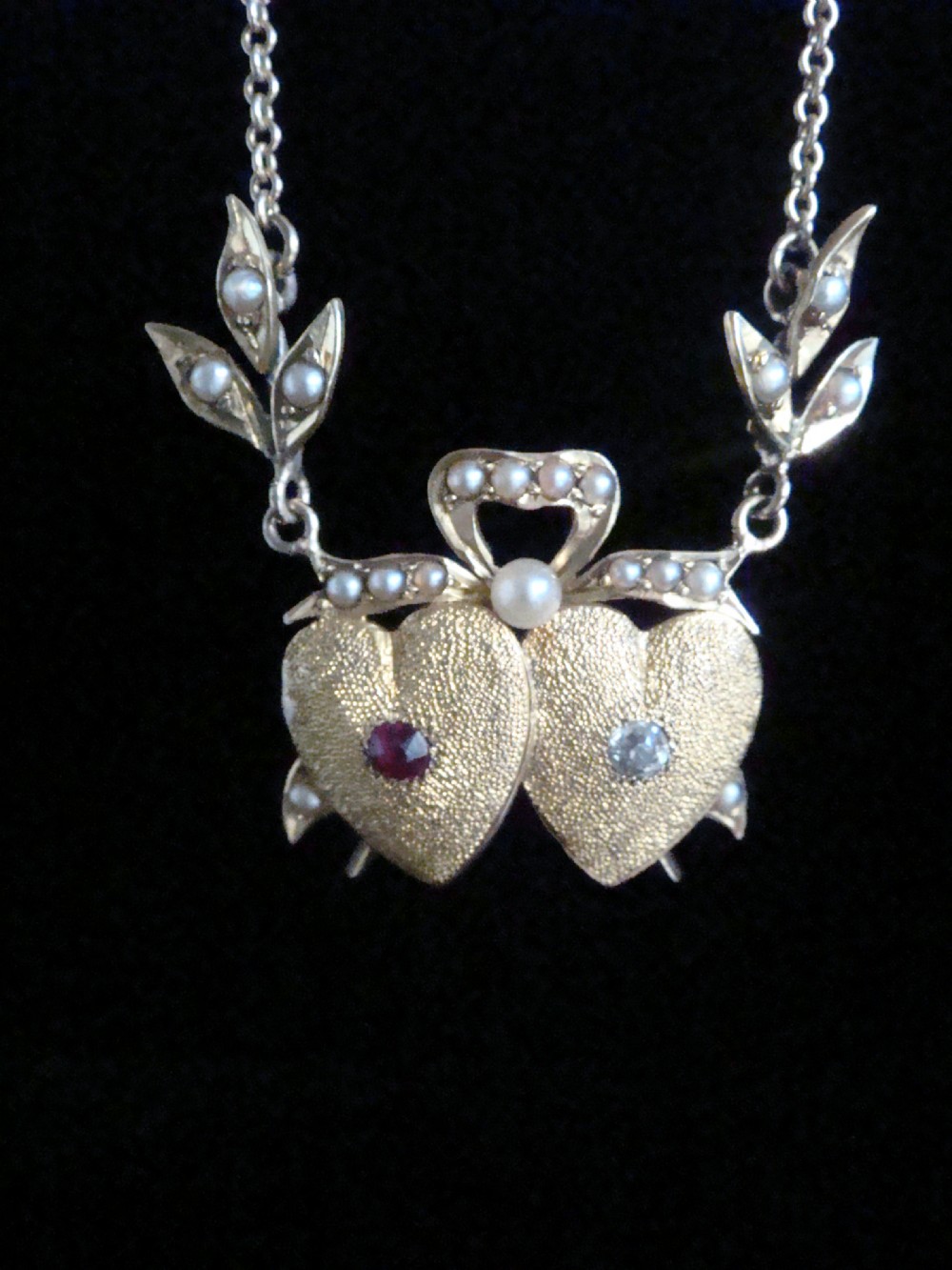 An Edwardian 15ct yellow gold double heart necklace featuring a central ruby, a rose cut diamond, and seed pearl accents on leaf motifs. View II
