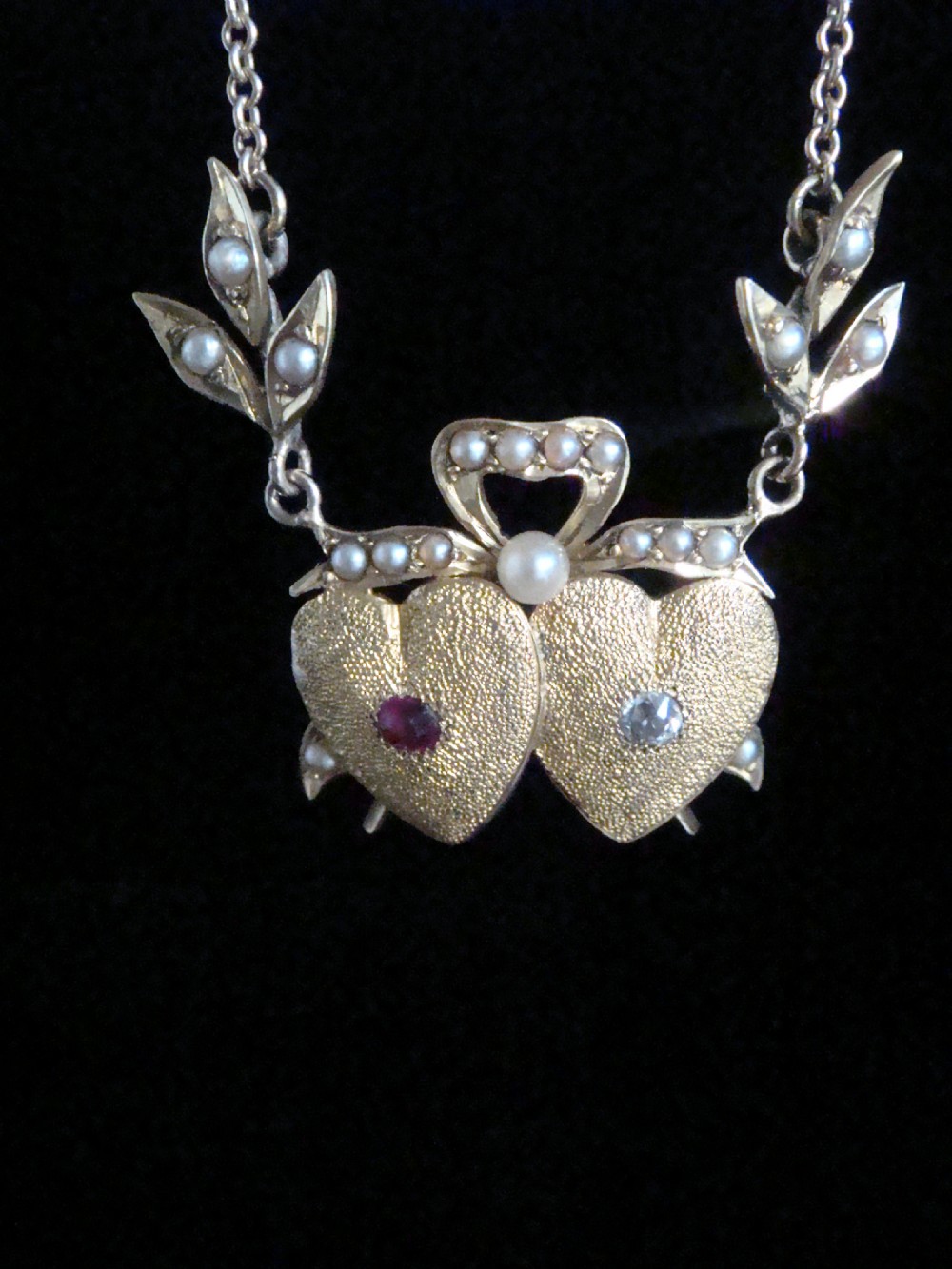 An Edwardian 15ct yellow gold double heart necklace featuring a central ruby, a rose cut diamond, and seed pearl accents on leaf motifs. View IV