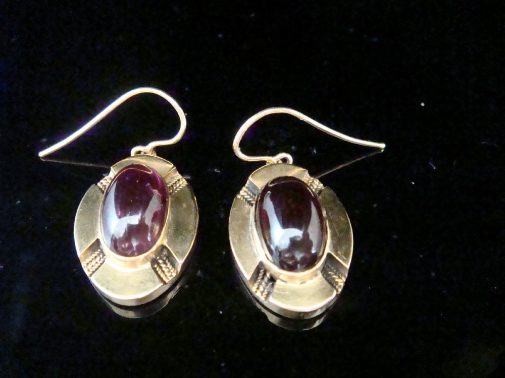A pair of Victorian 15ct gold drop earrings featuring oval cabochon garnets within textured golden frames with wire hooks.