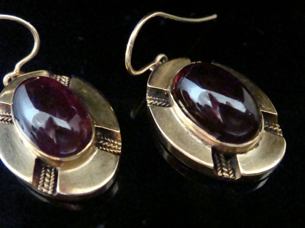 A pair of Victorian 15ct gold drop earrings featuring oval cabochon garnets within textured golden frames with wire hooks. View IV
