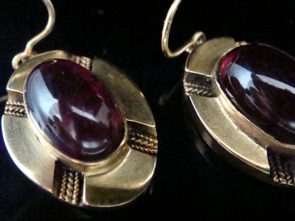 A pair of Victorian 15ct gold drop earrings featuring oval cabochon garnets within textured golden frames with wire hooks. View V