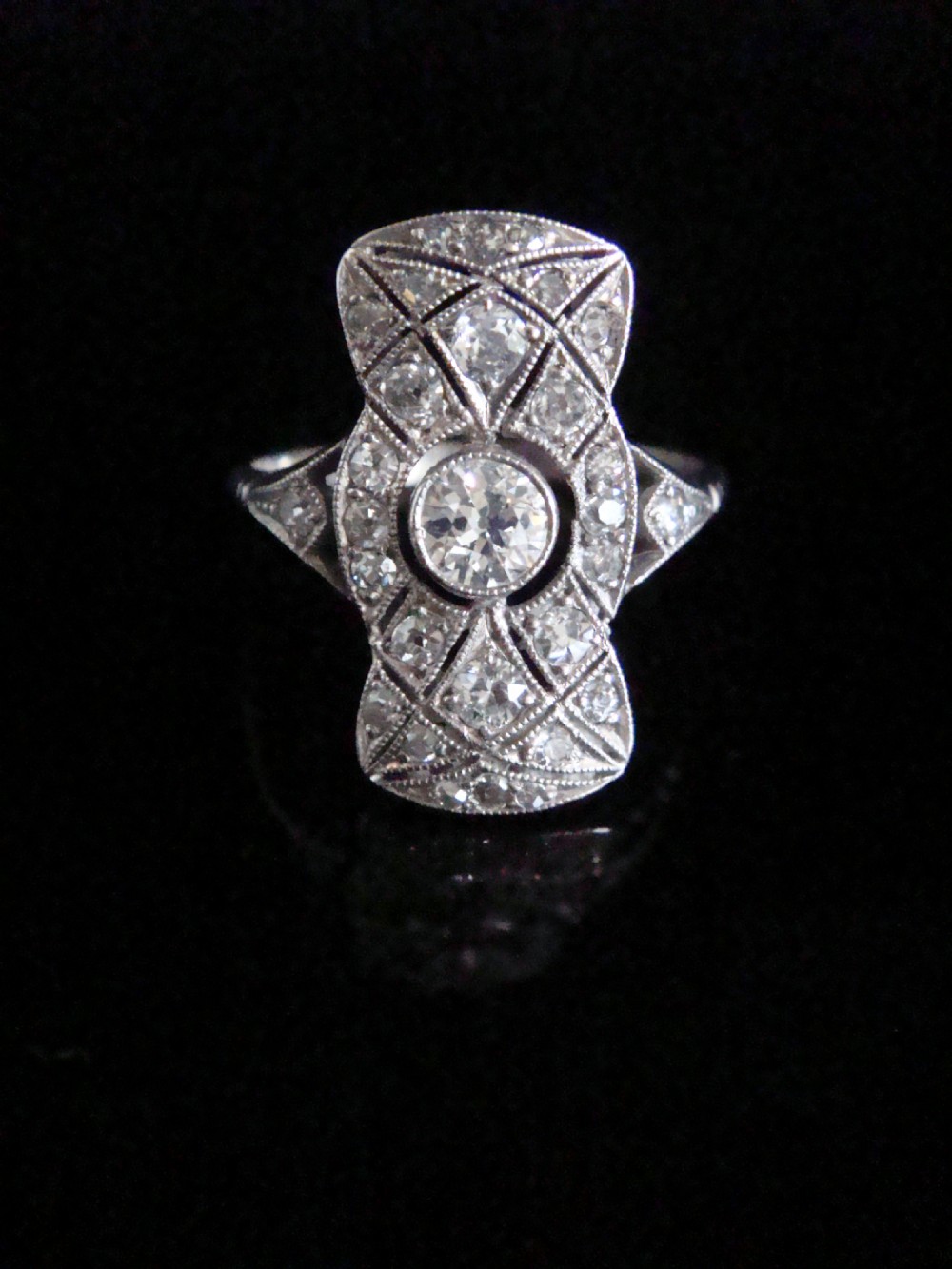 An Art Deco platinum plaque ring featuring a central round diamond set within an elongated geometric frame of smaller diamonds and millegrain detailing.