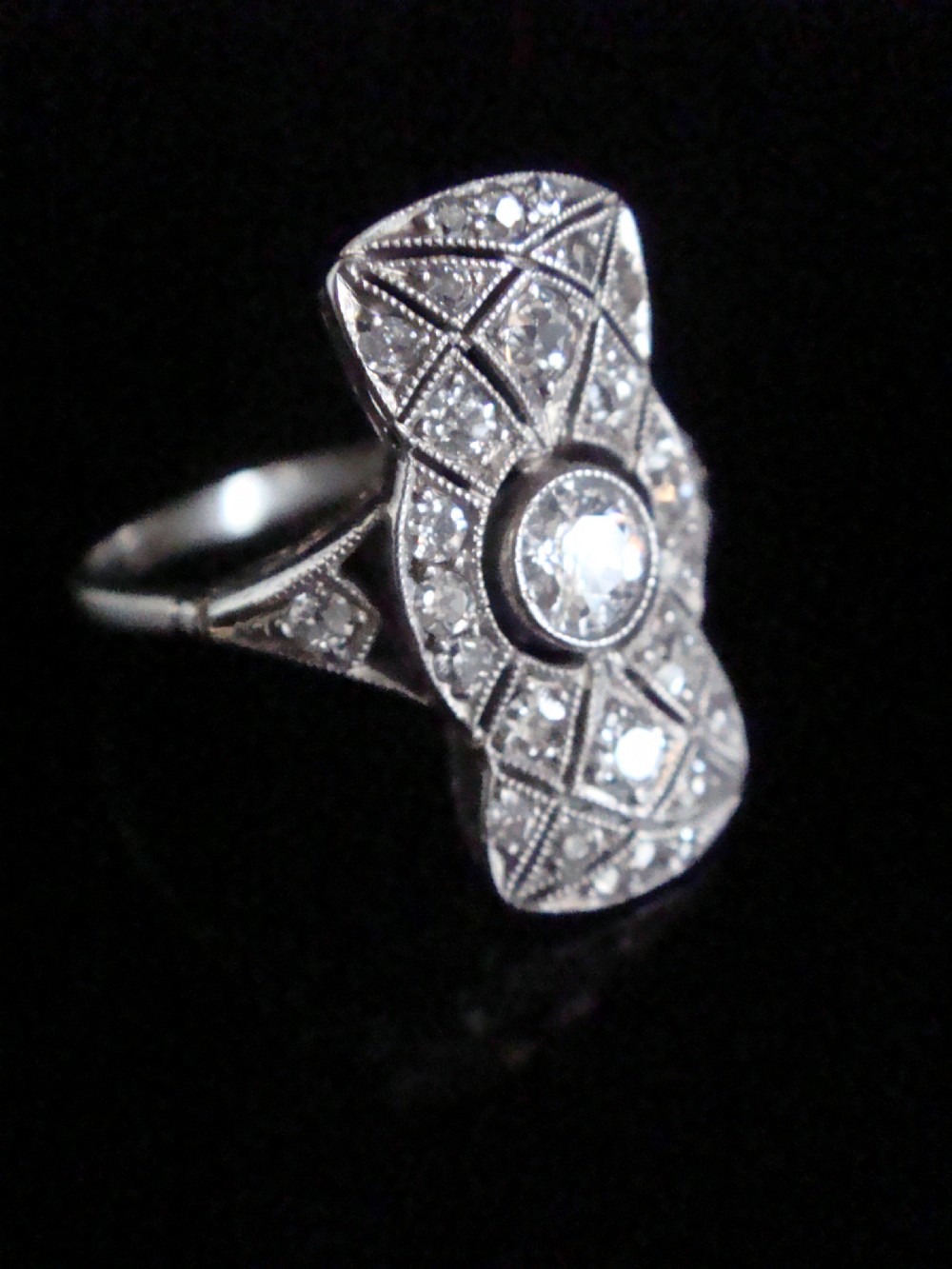 An Art Deco platinum plaque ring featuring a central round diamond set within an elongated geometric frame of smaller diamonds and millegrain detailing. View II