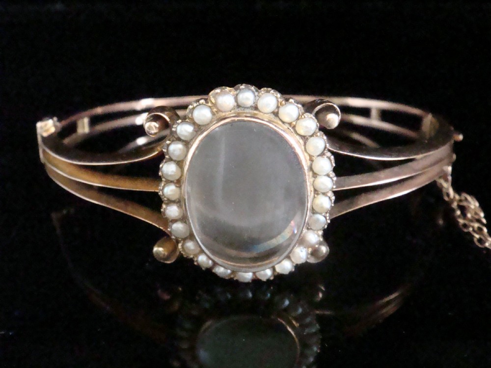 An antique 9ct yellow gold hinged bangle featuring a central oval glass shaker locket surrounded by a halo of small seed pearls.