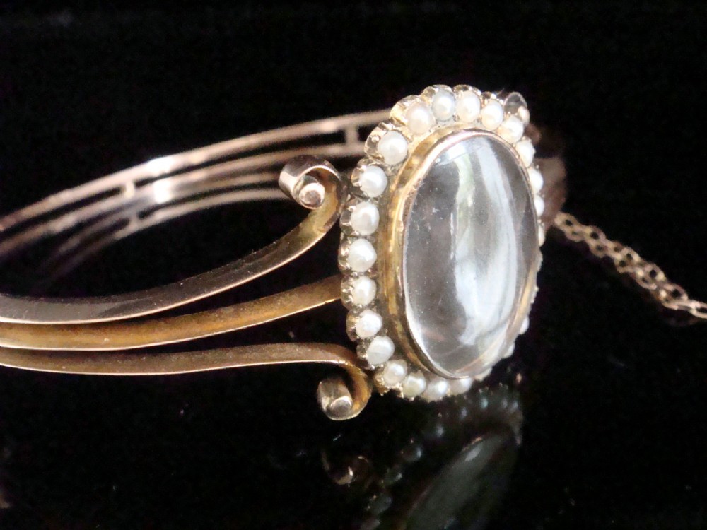 An antique 9ct yellow gold hinged bangle featuring a central oval glass shaker locket surrounded by a halo of small seed pearls. View II