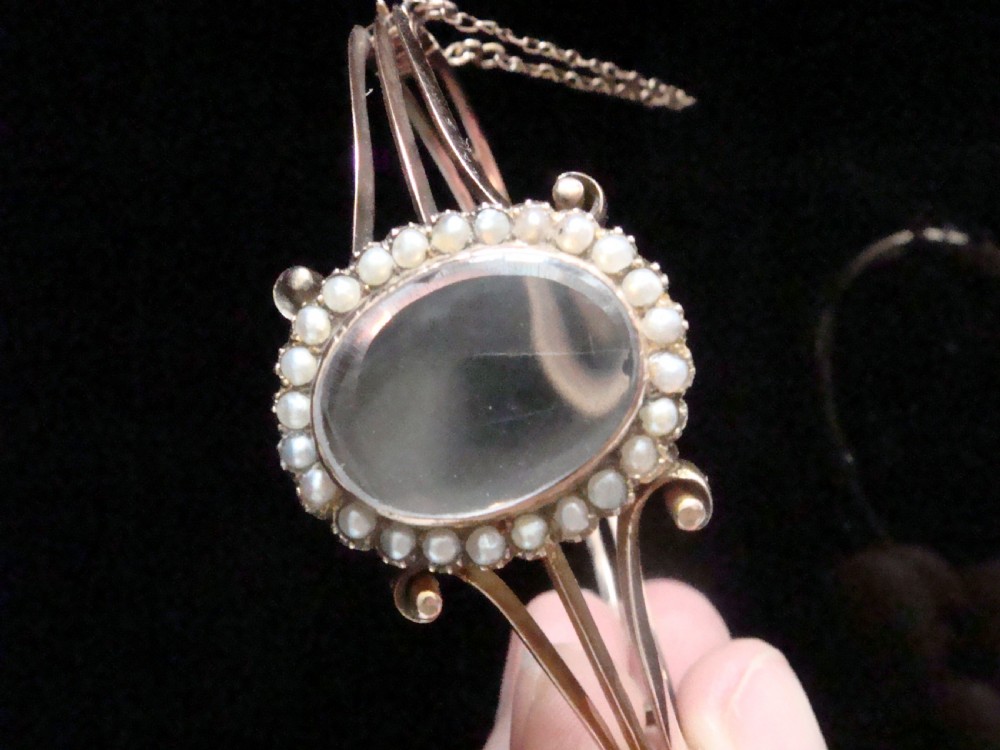 An antique 9ct yellow gold hinged bangle featuring a central oval glass shaker locket surrounded by a halo of small seed pearls. View IV