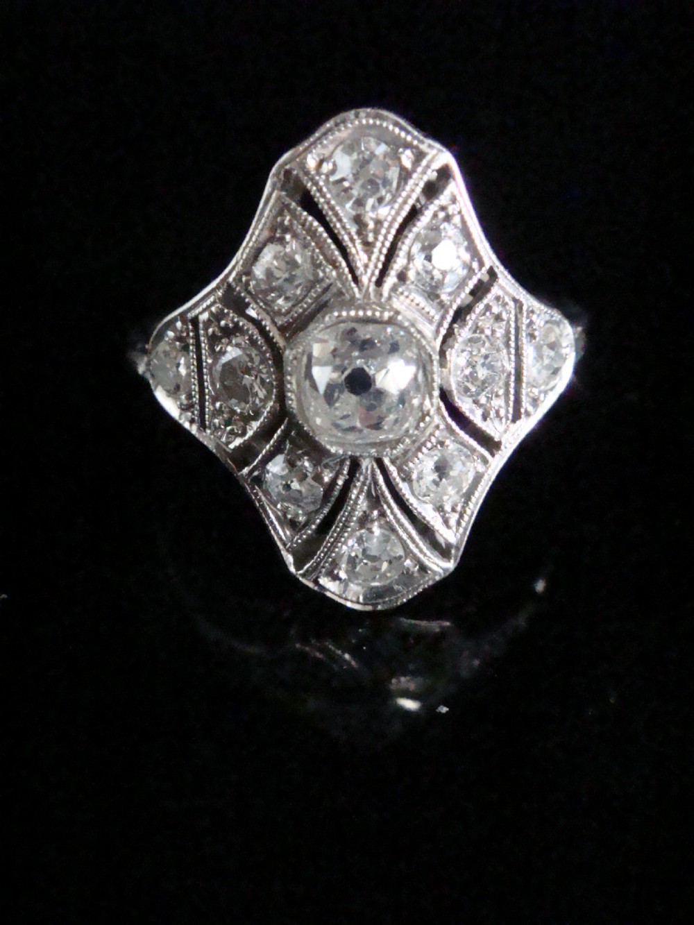 An Edwardian platinum dress ring featuring a central old cut diamond surrounded by geometric diamond-paved sections with millegrain edging.
