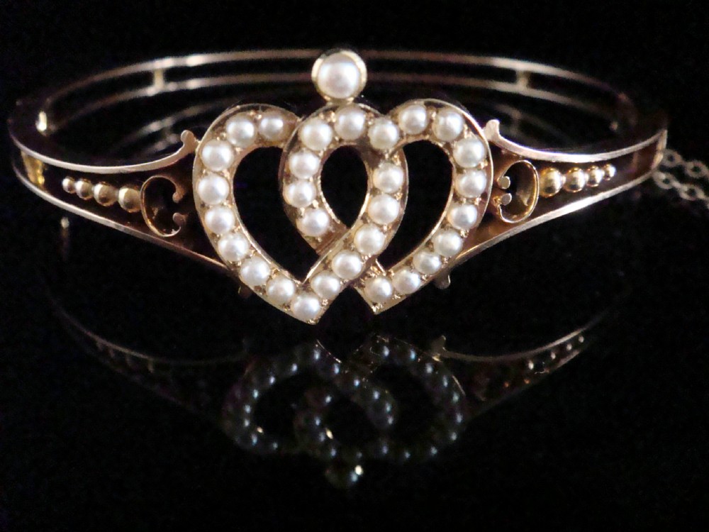 A 9ct gold hinged bangle featuring an intertwined double heart motif set with small seed pearls, complete with a safety chain against a dark reflective surface.
