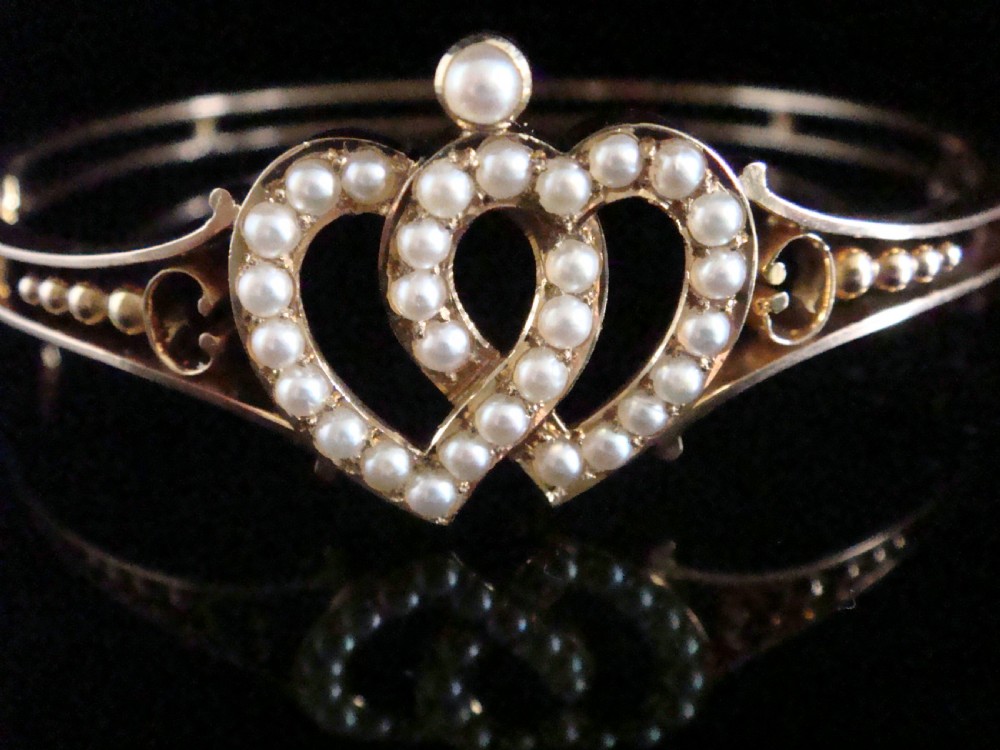 A 9ct gold hinged bangle featuring an intertwined double heart motif set with small seed pearls, complete with a safety chain against a dark reflective surface. View II
