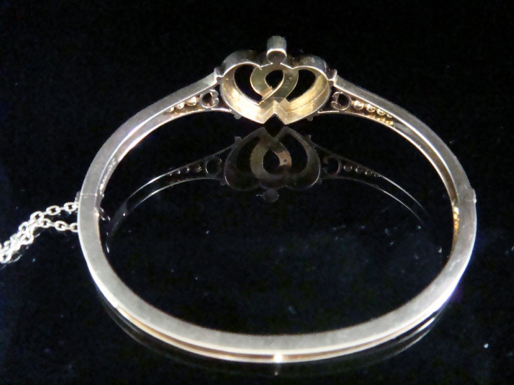 A 9ct gold hinged bangle featuring an intertwined double heart motif set with small seed pearls, complete with a safety chain against a dark reflective surface. View III