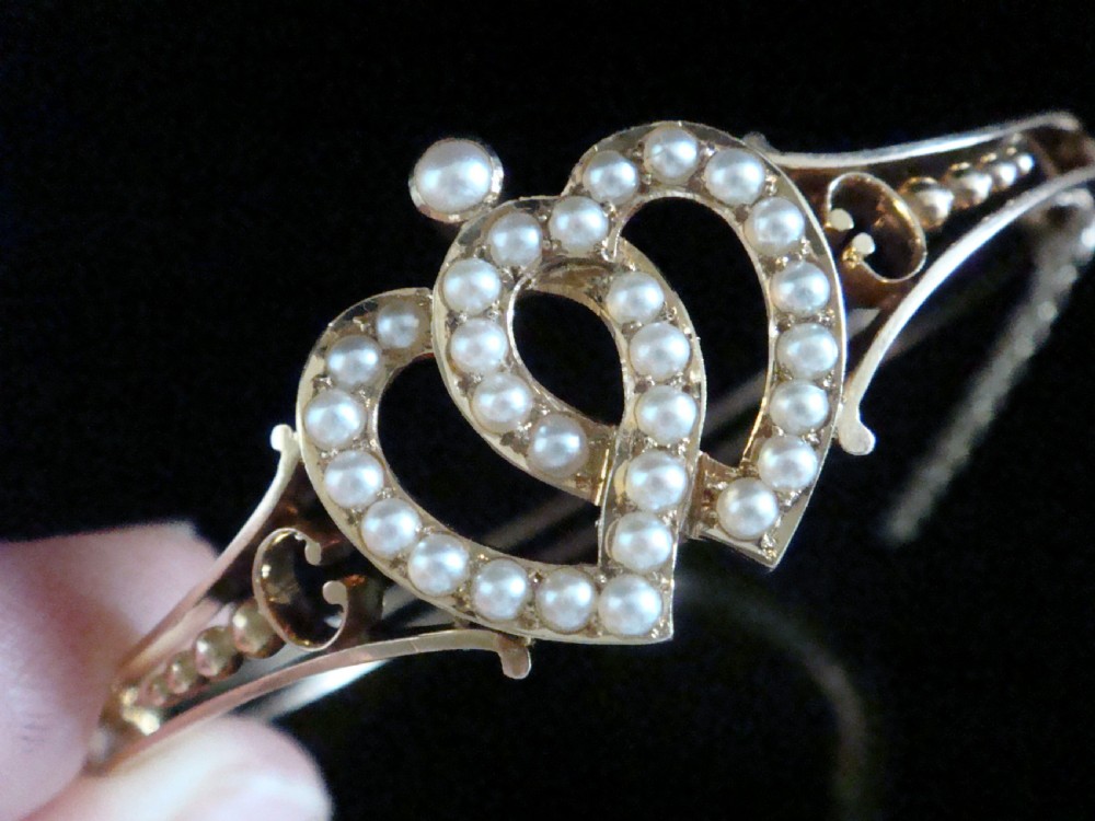 A 9ct gold hinged bangle featuring an intertwined double heart motif set with small seed pearls, complete with a safety chain against a dark reflective surface. View V