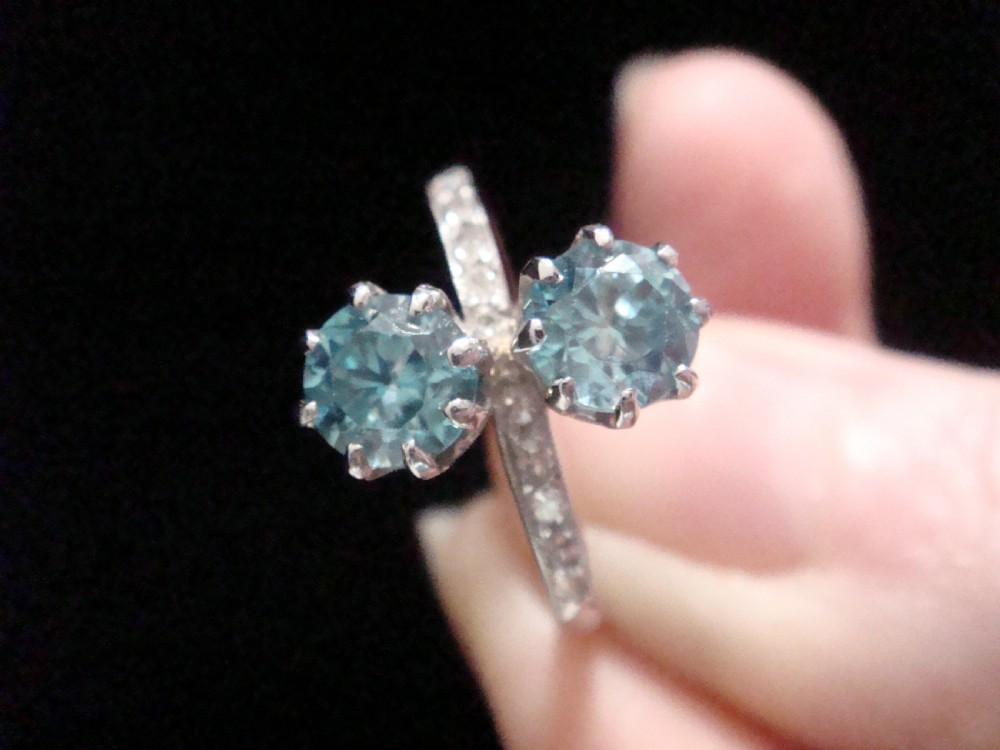 An Edwardian platinum ring featuring two round-cut blue zircon gemstones set vertically in a Toi et Moi design with diamond-set shoulders. View IV