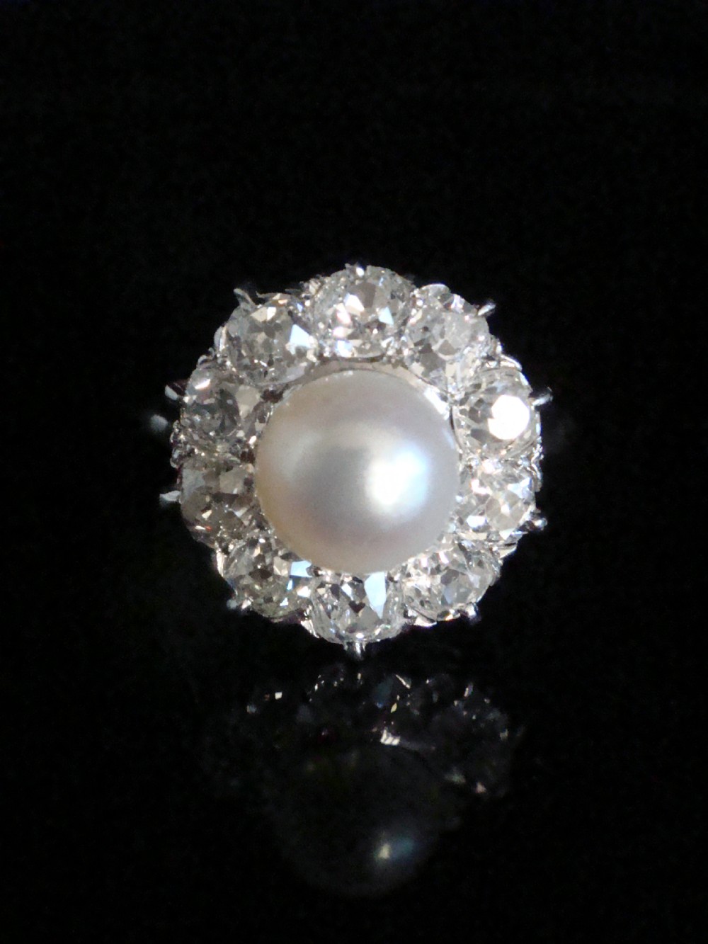 An Edwardian cluster ring featuring a central round pearl framed by a halo of old cut diamonds set in white metal.