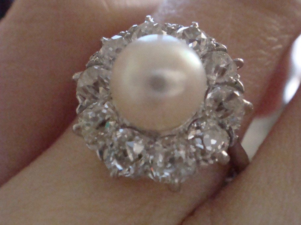An Edwardian cluster ring featuring a central round pearl framed by a halo of old cut diamonds set in white metal. View II