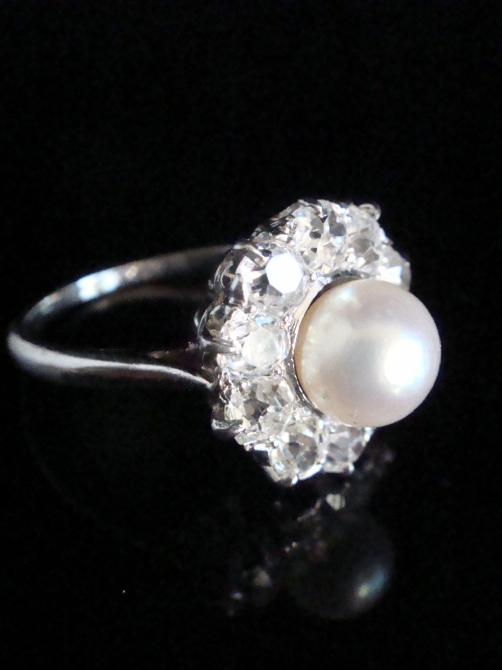 An Edwardian cluster ring featuring a central round pearl framed by a halo of old cut diamonds set in white metal. View V