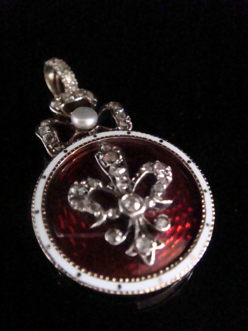 A circular Victorian gold locket featuring red guilloche enamel with a rose-cut diamond fleur-de-lis motif, surrounded by a white enamel border and topped with a diamond and pearl crown bail.