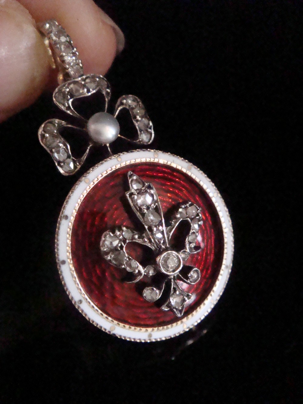 A circular Victorian gold locket featuring red guilloche enamel with a rose-cut diamond fleur-de-lis motif, surrounded by a white enamel border and topped with a diamond and pearl crown bail. View II