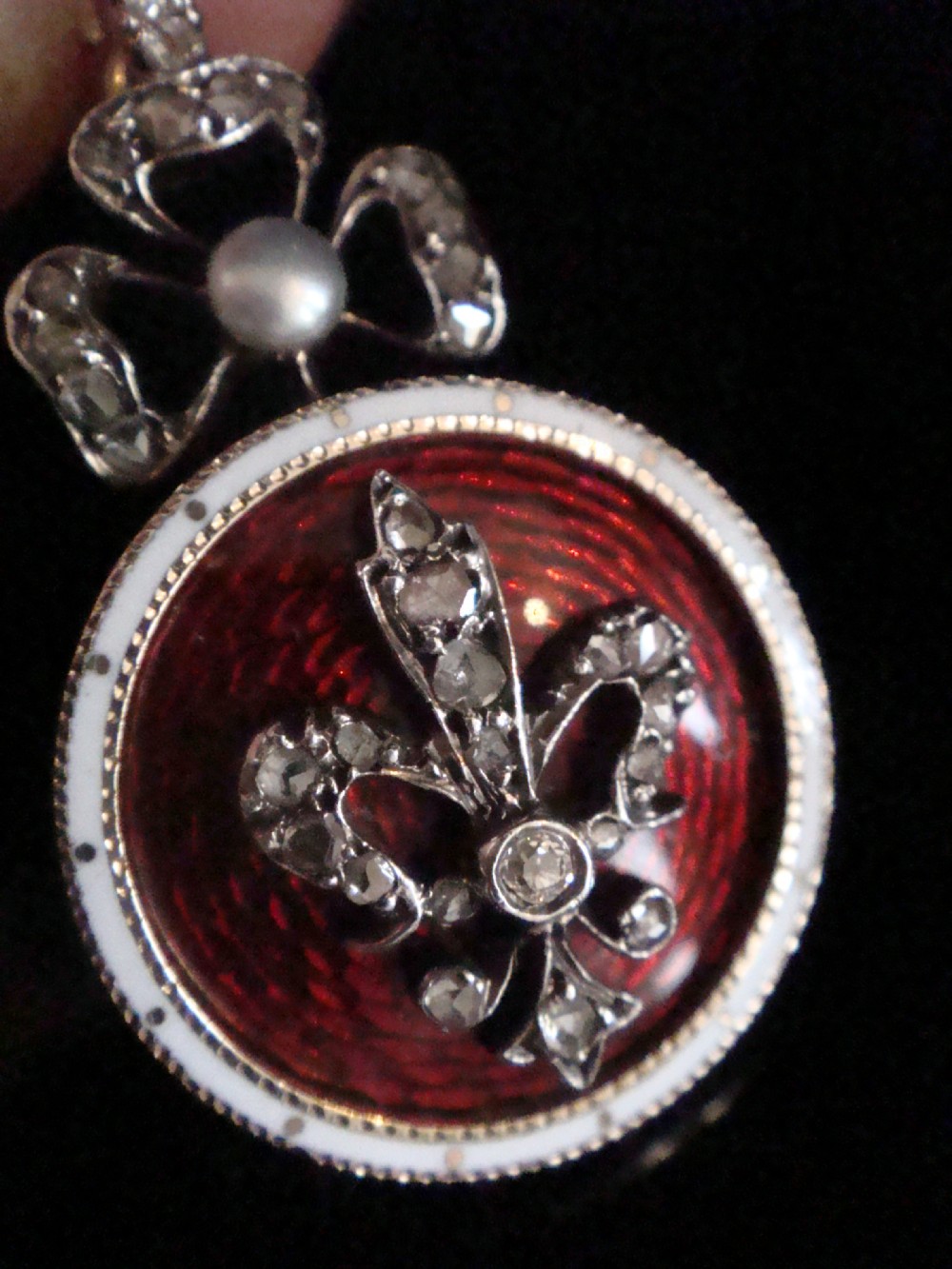A circular Victorian gold locket featuring red guilloche enamel with a rose-cut diamond fleur-de-lis motif, surrounded by a white enamel border and topped with a diamond and pearl crown bail. View III
