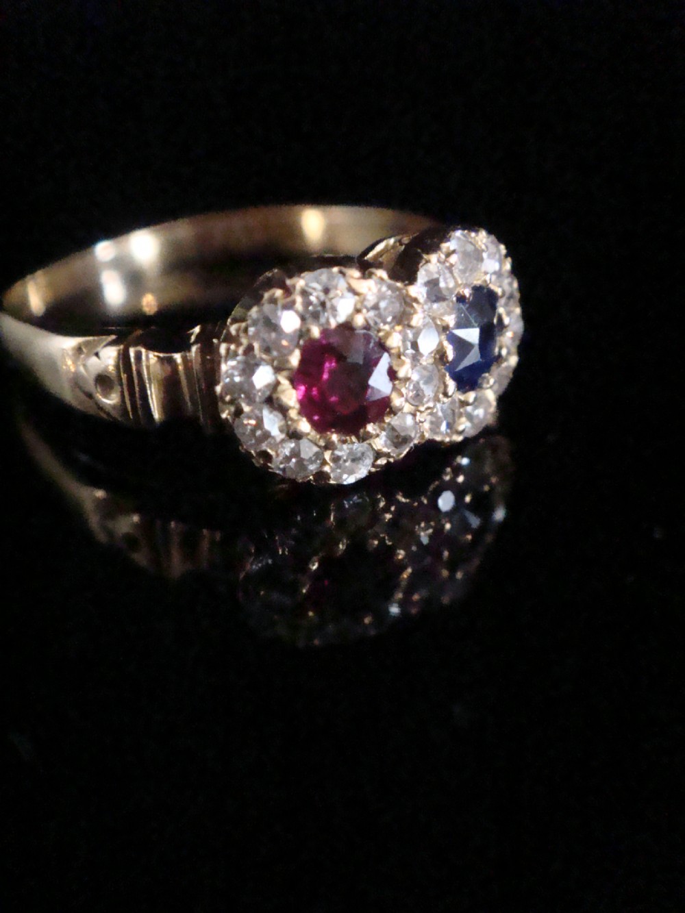 An Edwardian 18ct yellow gold Toi et Moi ring featuring a central ruby and sapphire, each encircled by a halo of old cut diamonds. View II