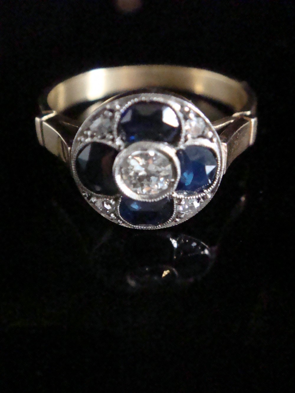 An Art Deco ring featuring a central round diamond surrounded by four dark blue sapphires arranged in a four-leaf clover motif with millegrain platinum borders and a gold shank.
