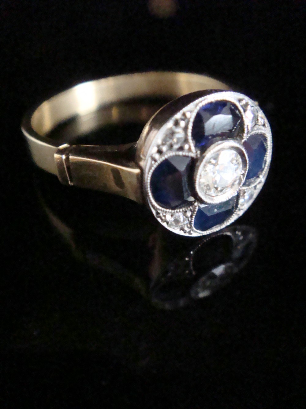 An Art Deco ring featuring a central round diamond surrounded by four dark blue sapphires arranged in a four-leaf clover motif with millegrain platinum borders and a gold shank. View II