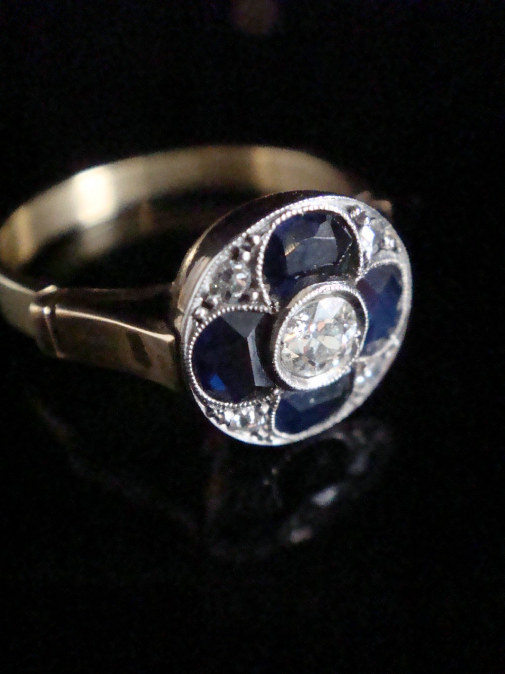 An Art Deco ring featuring a central round diamond surrounded by four dark blue sapphires arranged in a four-leaf clover motif with millegrain platinum borders and a gold shank. View III